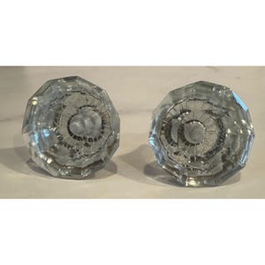 May include: Two clear glass door knobs featuring a multi-faceted, octagonal design. Each knob has a circular center with radiating lines extending toward the edges, creating a geometric pattern that reflects light through the transparent material.