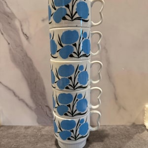 May include: A stack of five white ceramic mugs. Each mug features a blue floral design with black stems and leaves. The handles are a unique, three-lobed shape. The mugs are stacked vertically, showcasing the repeating pattern.