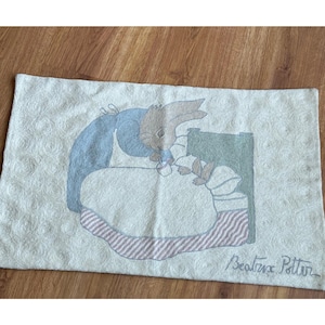 May include: A rectangular off-white textile featuring a textured swirl pattern and a central illustration of a brown rabbit in a blue dress feeding a smaller rabbit in a bed with a green headboard. The text Beatrix Potter is written in the corner.