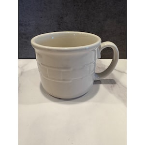 May include: A cream-colored ceramic mug featuring a raised, embossed basket-weave pattern around the exterior body. The mug has a wide, rounded bowl shape, a smooth interior, and a sturdy, curved handle attached to the side for gripping.