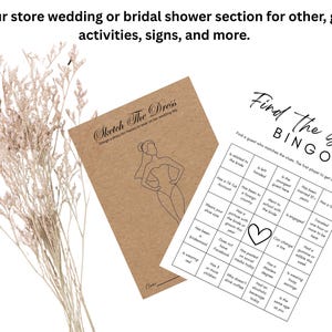 May include: A kraft paper card titled Sketch The Dress with a line drawing of a person in a veil, and a white Find The Guest Bingo game card with black text and a heart icon. Dried light beige flowers are positioned to the left of the cards.