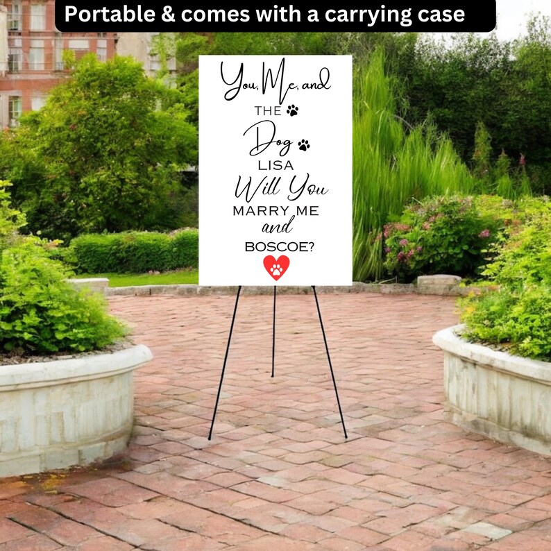 May include: A white sign on a black easel with the text "You, Me, and the Dog, Lisa, Will You Marry Me and Boscoe?" in black script. A red heart with a paw print is at the bottom. The sign is outdoors, surrounded by greenery.
