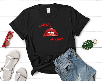 May include: Black t-shirt featuring a red graphic of a pair of lips with the text "antici...pation" printed on it. The t-shirt is laid out on a white wooden surface with a pair of silver sandals, a pair of sunglasses, and a green leaf keychain.