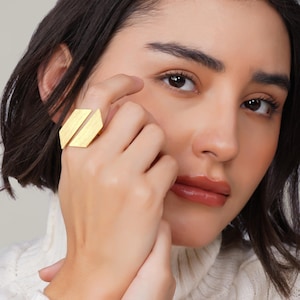 Può includere: Un anello color oro con un design geometrico caratterizzato da due forme rettangolari sfalsate. L'anello è indossato su un dito. Il design dell'anello è moderno e minimalista, con finitura in metallo spazzolato.