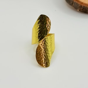Puede incluir: Un anillo de color dorado con un diseño cruzado de dos extremos curvos superpuestos. La superficie del metal tiene una textura martillada distintiva, creando una serie de pequeñas hendiduras reflectantes en todo el exterior visible.