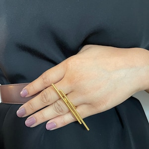 Op de afbeelding: Een goudkleurige ring met twee parallelle horizontale staven die zich uitstrekken over de breedte van twee vingers. De ring heeft een gladde, gepolijste metalen afwerking en een eenvoudig, lineair ontwerp dat plat op de vingers rust.