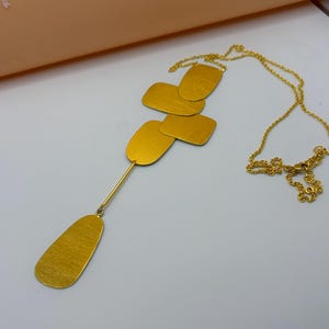 Pode incluir: Um colar dourado com uma corrente fina e um pendente composto por formas retangulares e ovais. O colar é apresentado numa superfície branca, destacando o seu design e cor. Os elementos do pendente são de cor dourada.