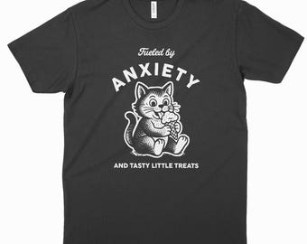 Peut inclure: Un t-shirt gris anthracite à manches courtes avec un motif blanc représentant un chat mangeant un cornet de glace. Le texte au-dessus du chat indique Fueled by ANXIETY et le texte en dessous indique AND TASTY LITTLE TREATS en lettres blanches.