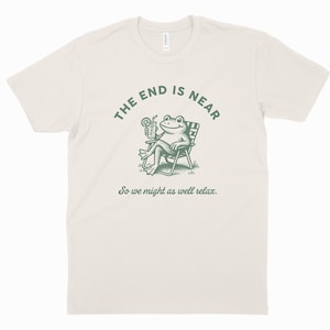 May include: Off-white t-shirt featuring a green illustration of a frog in a deckchair, holding a cocktail. The text "THE END IS NEAR" is above the frog, with "So we might as well relax." underneath.