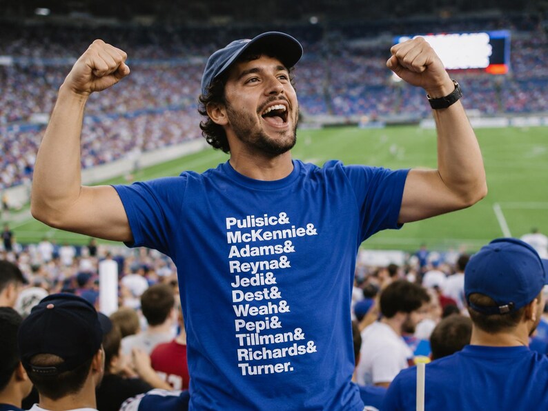 May include: A royal blue short-sleeved t-shirt featuring a vertical list of white printed names including Pulisic, McKennie, Adams, Reyna, Jedi, Dest, Weah, Pepi, Tillman, Richards, and Turner, each separated by an ampersand symbol.