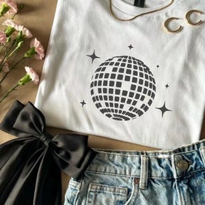 May include: White t-shirt featuring a black disco ball design and star shapes. The image includes denim shorts, a black bow, gold jewellery, and pink flowers. The items are arranged on a neutral surface.