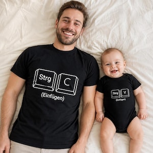 May include: A black short-sleeved t-shirt and a black short-sleeved baby bodysuit. The t-shirt features white keyboard key graphics labeled Strg and C with the text Einfügen. The bodysuit features white keyboard key graphics labeled Strg and V.