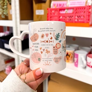 Könnte beinhalten: Weiße Keramiktasse mit herzförmigem Henkel, verziert mit inspirierenden Sätzen und floralen Illustrationen in Pfirsich und Rosa. Die Tasse enthält den Text "Growth takes time. Be patient with yourself."