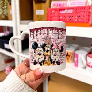 Peut inclure: Tasse en céramique blanche avec une anse en forme de cœur. La tasse présente un motif avec Mickey Mouse, Dingo et Minnie Mouse, avec un château rose et des accents dégoulinants. Les mots "I Love You" sont également visibles.