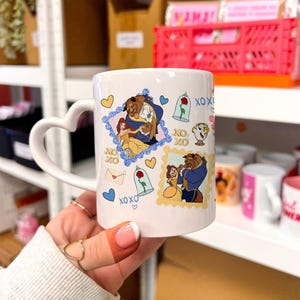 Op de afbeelding: Witte keramische mok met een hartvormig handvat, met illustraties van Belle en het Beest-personages. De mok heeft een herhalend patroon van harten, rozen en de letters "XOXO".