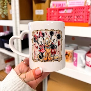 Peut inclure: Tasse en céramique blanche avec une anse en forme de cœur. La tasse présente un graphisme coloré de Mickey Mouse, Minnie Mouse et Pluto, entourés de cœurs et d'une bordure léopard. La tasse est de taille standard pour le café ou le thé.