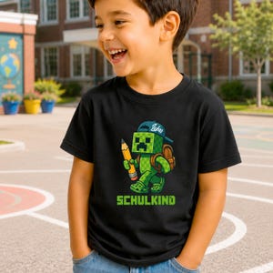 Könnte beinhalten: Ein schwarzes Kurzarm-T-Shirt mit einer Grafik eines grünen, verpixelten Charakters, der eine blaue Kappe mit dem Namen Lukas trägt, einen braunen Rucksack aufhat und einen gelben Stift hält. Der Text SCHULKIND steht in Grün unter der Figur.