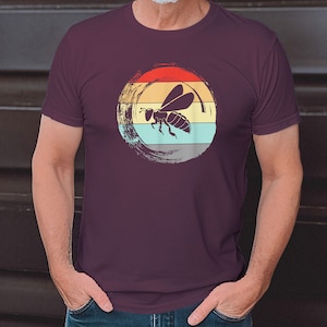 Könnte beinhalten: Ein maroonfarbenes T-Shirt mit einer Retro-Grafik einer Biene, die in einem Kreis mit einem Sonnenuntergangs-Hintergrund fliegt.