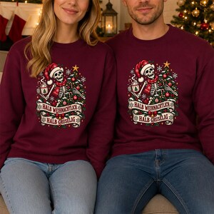 Puede incluir: Una sudadera de manga larga color burdeos con un gráfico de un esqueleto con gorro y bufanda de Papá Noel, junto a un árbol de Navidad decorado. El diseño incluye una pancarta con el texto "HALB WEIHNACHTLICH HALB GRUSELIG".