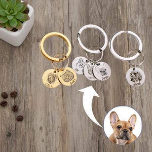 Può includere: Portachiavi con targhette identificative personalizzate per animali domestici. Le targhette rotonde presentano ritratti di animali domestici e nomi incisi. I portachiavi sono dorati e argentati. Sfondo in legno con una pianta e chicchi di caffè.