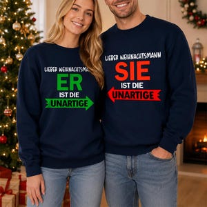 Op de afbeelding: Twee marineblauwe sweaters met ronde hals en witte, groene en rode tekst. De linker sweater toont LIEBER WEIHNACHTSMANN ER IST DIE UNARTIGE met een groene pijl. De rechter sweater toont LIEBER WEIHNACHTSMANN SIE IST DIE UNARTIGE met een rode pijl.