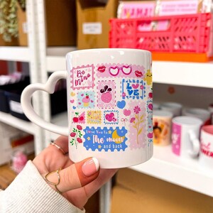 Puede incluir: Taza de cerámica blanca con asa en forma de corazón, decorada con coloridos diseños de sellos postales. Los sellos presentan corazones, labios y el texto "Be mine" y "Love You to the moon and back".