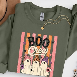 Peut inclure: Un sweat-shirt à col rond vert olive avec une impression graphique de trois fantômes portant des bonnets colorés. Le texte indique BOO Crew. Le motif comprend des rayures verticales roses et orange, des petites étoiles et deux tasses.