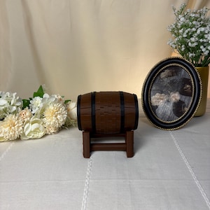 May include: A miniature brown barrel with black bands, resting on a small brown stand. The barrel has a woven texture. A decorative oval frame with a black and gold border and a vase of white flowers are in the background.