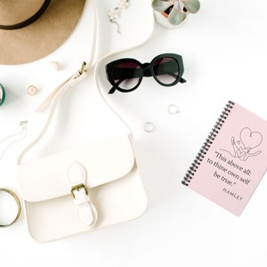 Puede incluir: Una libreta de espiral rosa con una cita de Hamlet: "Sobre todo: sé fiel a ti mismo." La cita está escrita en texto negro y está acompañada por un dibujo lineal de dos manos sosteniendo un corazón.