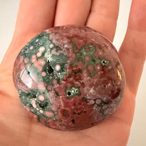 May include: A polished, rounded gemstone with a mix of colors. The stone features a base of deep red with patches of green and pink, creating a speckled appearance. The surface is smooth and reflective.