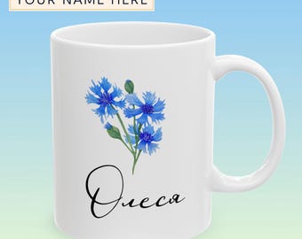 Op de afbeelding: Witte keramische mok met een aquarel illustratie van blauwe korenbloemen en de naam "Олеся" in zwart schrift. De mok heeft een gebogen handvat en de tekst "YOUR NAME HERE" boven de mok.