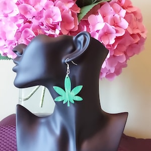 Könnte beinhalten: Grüne Marihuana-Blatt-Ohrringe an einem schwarzen Mannequin-Kopf. Die Ohrringe sind aus Holz und haben einen silbernen Haken.