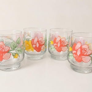 May include: Four clear glass tumblers with a floral design. Each tumbler features a large red rose with yellow and pink accents, surrounded by green leaves. The glasses are set against a white background.
