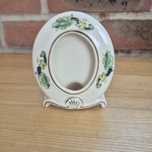 May include: An oval-shaped ceramic picture frame in an off-white colour. The frame features a decorative green and yellow floral pattern along the sides and top. A gold-coloured trim outlines the inner oval and the base, which includes a shell motif.
