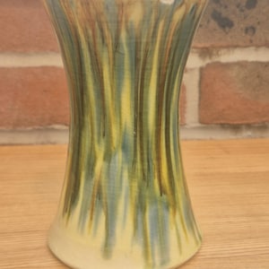 May include: A ceramic vase with a flared top and base, featuring a cream-colored glaze with vertical drip patterns in shades of green, blue, and brown. The surface has a visible crackle finish and a small chip on the upper rim edge.