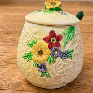 May include: A ceramic jar with a lid featuring a yellow honeycomb-textured surface. The jar is decorated with raised, colorful floral motifs in red, yellow, and purple, with green leaves. A small green handle is attached to the lid.