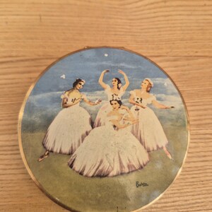 May include: A round metal compact case featuring a colorful illustration of four dancers in white dresses on the lid. The artwork depicts the figures against a blue sky and green ground, with the signature Baron visible in the bottom right corner.