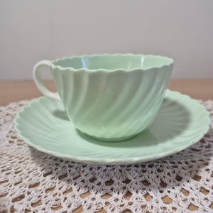 May include: A pale mint green teacup and matching saucer set featuring a delicate swirl pattern throughout the body. The cup has a curved handle and a scalloped rim, while the saucer also displays a matching scalloped edge design.