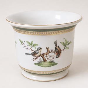 Puede incluir: Una maceta de porcelana blanca con una escena pintada a mano de dos pájaros pequeños posados en una rama marrón con hojas verdes. El recipiente presenta una banda verde oscuro y un borde dorado en la parte superior e inferior del objeto.