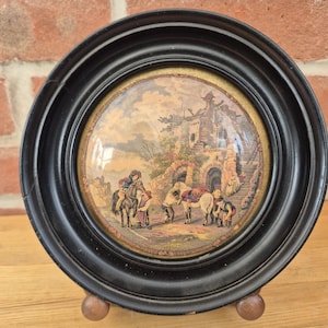 May include: A circular wall plaque featuring a convex glass cover set within a black frame. The print depicts a pastoral scene with figures, horses, and stone ruins under a cloudy sky. The piece is supported by two small, rounded wooden feet.