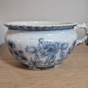 May include: A white ceramic chamber pot featuring a dark blue floral pattern around the exterior. The vessel has a wide, rounded rim with a scalloped edge, a curved handle on the right side, and a smooth, glossy finish throughout the piece.