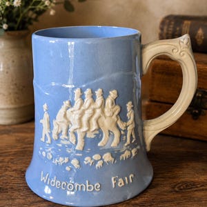 Può includere: Tazza in ceramica azzurra con manico color crema. Presenta una scena in bassorilievo bianca di persone a cavallo e la scritta "Widecombe Fair". La tazza ha un design classico ed è probabilmente vintage.