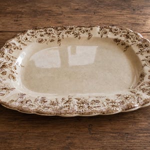 Pode incluir: Uma travessa oval antiga com um centro creme e uma borda floral marrom. A borda tem uma borda ondulada. A travessa é feita de cerâmica e fica em uma superfície de madeira. O padrão floral inclui pequenas flores semelhantes a margaridas.