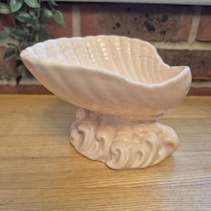May include: A pale pink ceramic bowl shaped like a large seashell, featuring a ribbed interior texture and a base sculpted to resemble stylized ocean waves. The glossy finish reflects light across the curved surface of the decorative vessel.