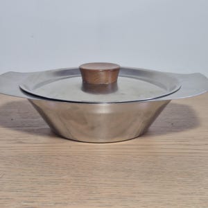 May include: A stainless steel serving bowl with a matching lid featuring a circular wooden knob handle. The bowl has wide, flat side handles integrated into the rim, creating a sleek, low-profile design with a polished metallic finish.