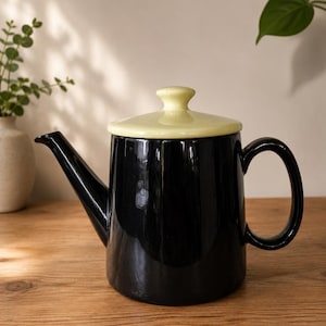 May include: A black ceramic teapot with a pale yellow lid and handle. The teapot has a long spout and sits on a wooden surface. The background is a neutral colour with a plant in the corner.