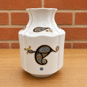 Può includere: Vaso in ceramica bianca dalla forma geometrica unica. Il vaso presenta motivi colorati a forma di paisley in blu, arancione e giallo. Rifiniture dorate accentuano il bordo. Un elemento decorativo per la casa.