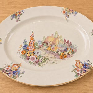 Peut inclure: Plat ovale en céramique crème. Il présente une image centrale d'un cottage et d'un jardin, entourée de grappes florales. Le bord est orné d'une bordure dorée. Environ 30 cm de long.