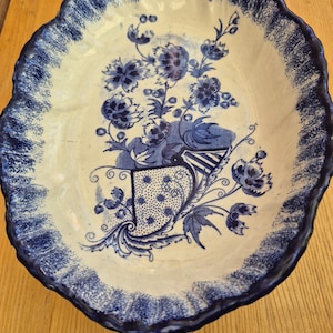 May include: An oval ceramic bowl with a scalloped edge featuring a blue and white floral pattern. The centre displays a cornucopia overflowing with flowers, while the rim is decorated with a dark blue sponged or brushed texture design.