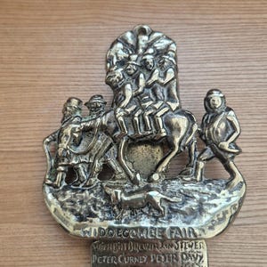 May include: A brass-colored metal plaque depicting a scene of people riding a horse, with a dog running alongside. The plaque has the words "WIDDECOMBE FAIR" and other text engraved on it. The plaque is mounted on a wooden surface.